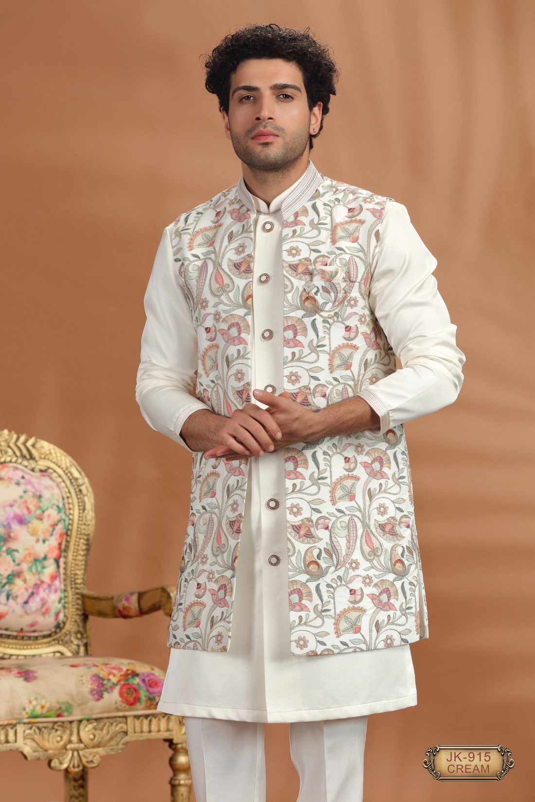 Man wearing cream sherwani with floral embroidery, standing beside ornate chair, traditional Indian wedding outfit.