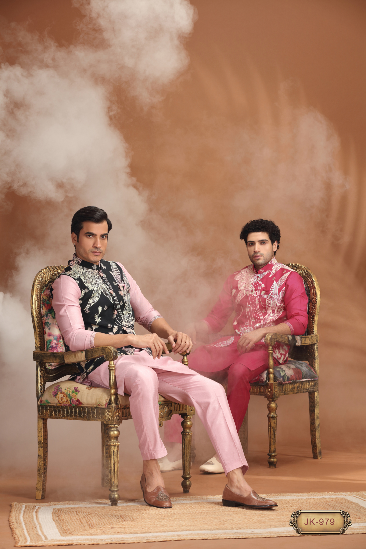 Two men in embroidered pink and red traditional South Asian sherwanis, seated on antique floral chairs with smoke effect.